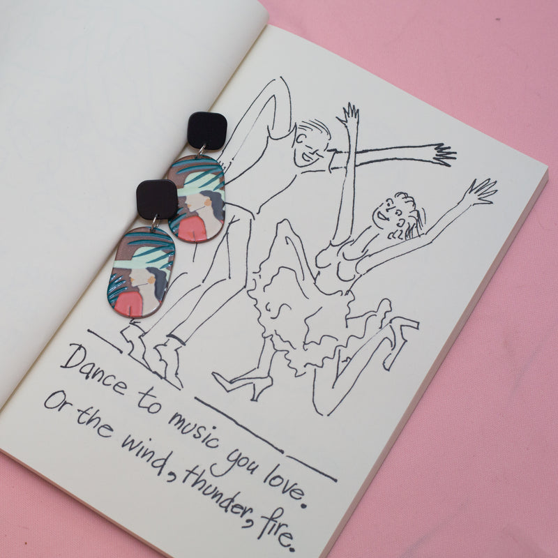 Studio Joy earrings with a woman among palm leaves on a book with an illustration of people dancing